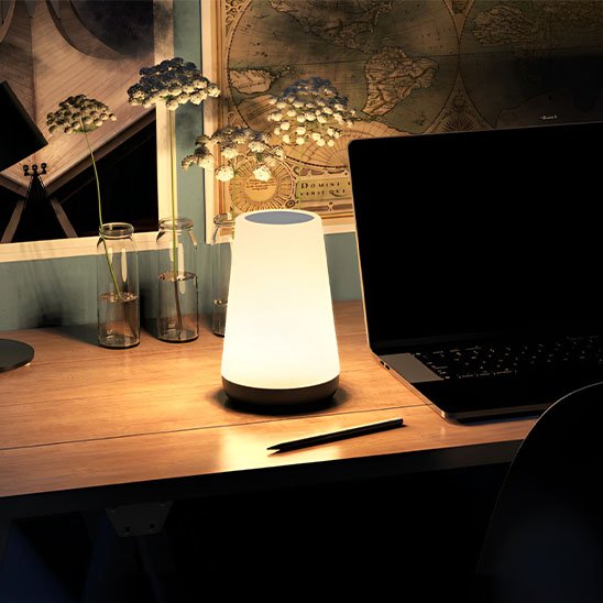 Sur un bureau à côté d'un ordinateur portable on voit la Lampe de chevet tactile | LumiTouch allumée.