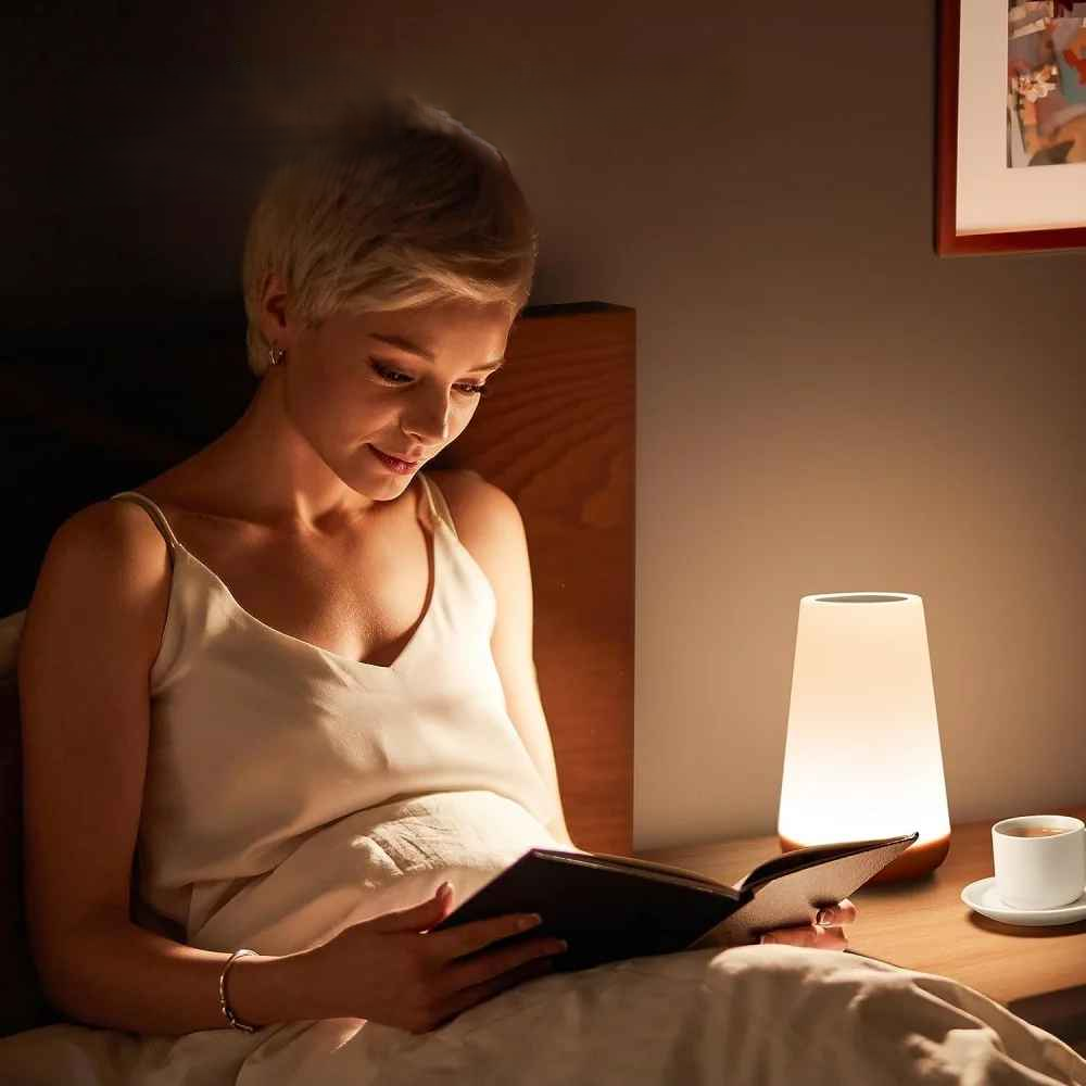 On voit une femme assise dans son lit avec un livre et la Lampe de chevet tactile | LumiTouch, sur sa table de chevet.