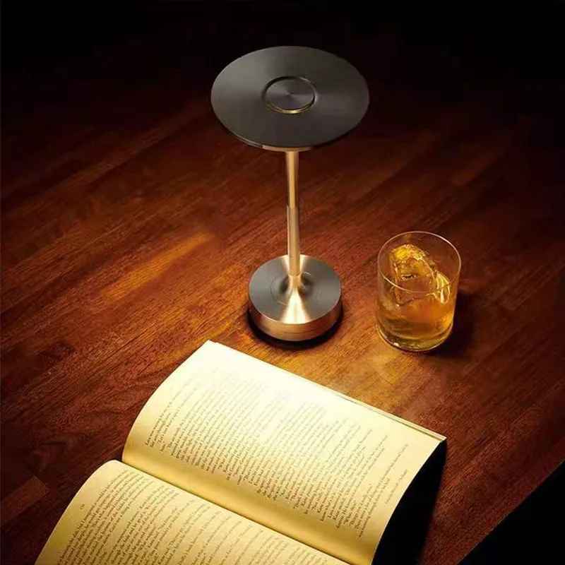 Lampe MoedernTouch de couleur argent posée sur une table près d'un livre ouvert avec un verre contenant une boisson avec de la glace