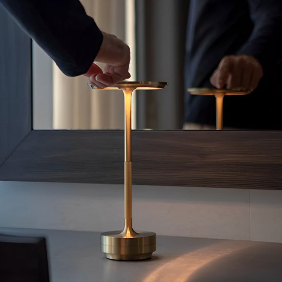 L'image montre une main d'homme qui appuie sur le dessus de la Lampe MoedernTouch de couleur or posée sur table devant un miroir dans lequel on voit le reflet de l'homme.