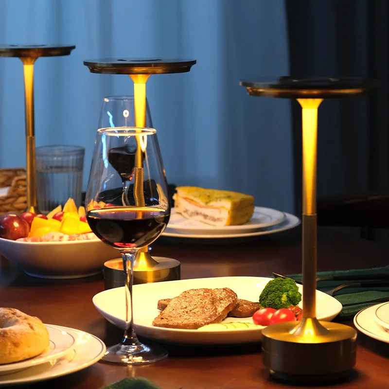 3 Lampes MoedernTouch de couleur or posées sur table On y voit des assistes et des bolles remplies de nourriture, des verre de vin rouge et un verre d'eau