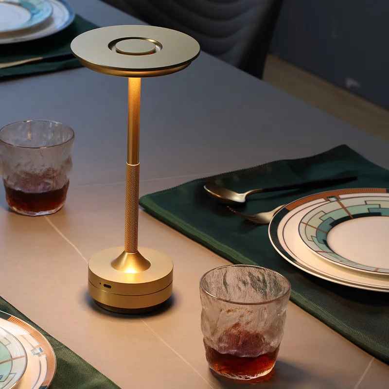 Lampe MoedernTouch de couleur or posée sur une table mise pour les convives et deux verre contenant une boisson qui sont posée de chaque côté de la lampe