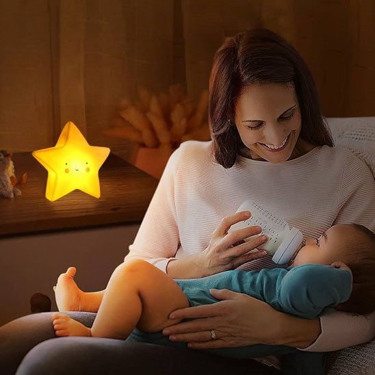 Une veilleuse bébé | Étoile Douce Nuit de couleur Jaune posée sur un meuble éclaire une maman qui donne le biberon à son bébé