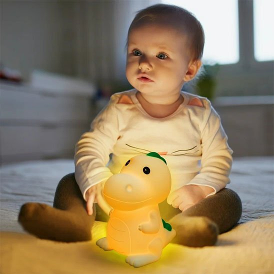 Un bébé est assis sur un lit avec la veilleuse bébé | dinosaure DinoNuit devant lui