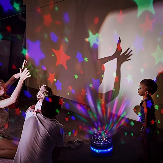 On voit deux jeunes enfants dont une fillette dans les bras du papa. La maman fait aller ses mains, comme les ailes d'un oiseau, dont l'ombre se voit au mur au travers des étoiles multicolores émises par la veilleuse projection plafond | CielEtoileMagique.
