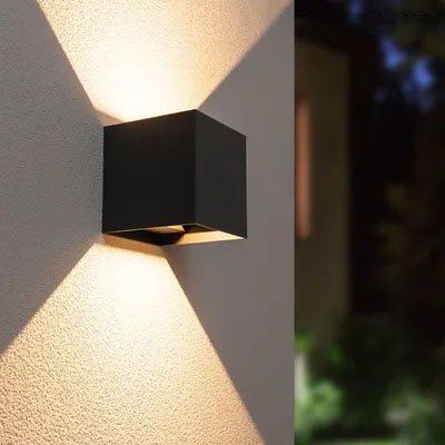 On voit l'applique murale pour extérieur | Luminesence : de 6 ou 12 watts sur le mur près du coin d'une maison. Elle est de couleur noire, de forme cubique. Ses dimensions sont de 10 cm de large par 10 cm de haut et 10 cm de profond. Elle diffuse de la lumière directe sur le mur vers le haut et le bas.