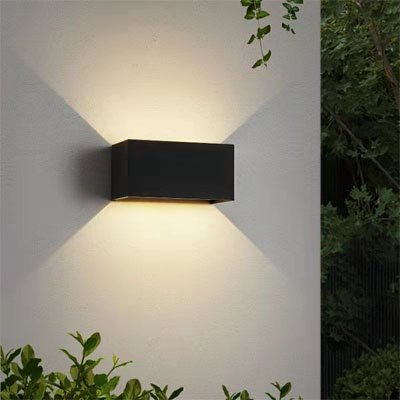 On voit l'applique murale pour extérieur | Luminesence : de 24 watts installée sur le mur près du coin d'une maison. Elle est de couleur noire, de forme rectangulaire. Ses dimensions sont de 20 cm de large par 10 cm de haut et 10 cm de profond. Elle diffuse de la lumière directe sur le mur vers le haut et le bas. Elle comprend des déflecteurs ajustables de 0 à 120 degrés, pour obtenir des effets lumineux personnalisables comme vous le souhaitez.