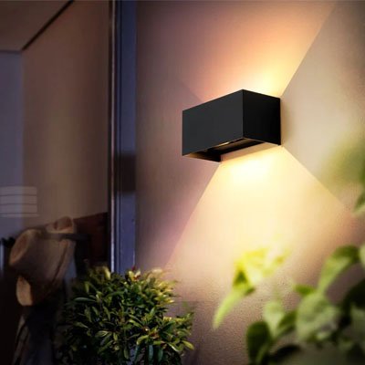 On voit l'applique murale pour extérieur | Luminesence : de 24 watts installée sur lu mur près d'une grande vitre d'entrée. l'applique murale de couleur noire se distingue par sa forme rectangulaire. Ses dimensions sont de 20 cm de large par 10 cm de haut et 10 cm de profond. Elle diffuse de la lumière directe sur le mur vers le haut et le bas.