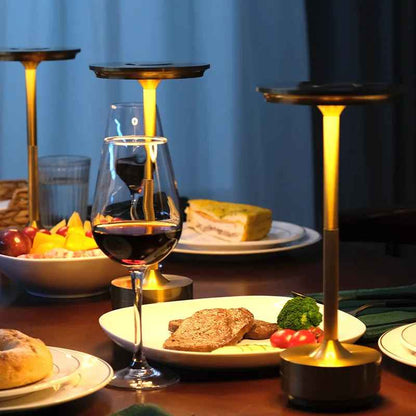 3 Lampes MoedernTouch de couleur or posées sur table On y voit des assistes et des bolles remplies de nourriture, des verre de vin rouge et un verre d'eau
