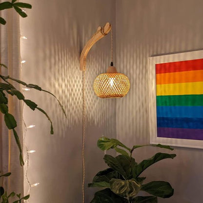 On voit la lueur texturée sur le mur  auquel est accroché l'applique murale rotin | Artisanale, qui pend au-dessus d'une grosse plante verte. On voit ce même éclairage projeté sur le cadre sur le mur adjacent